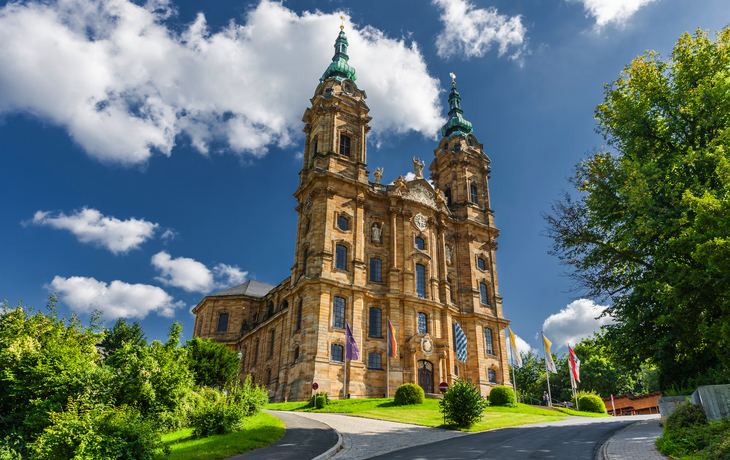 Barocke Kirche mit zwei Türmen vor blauem Himmel und grünen Bäumen.
